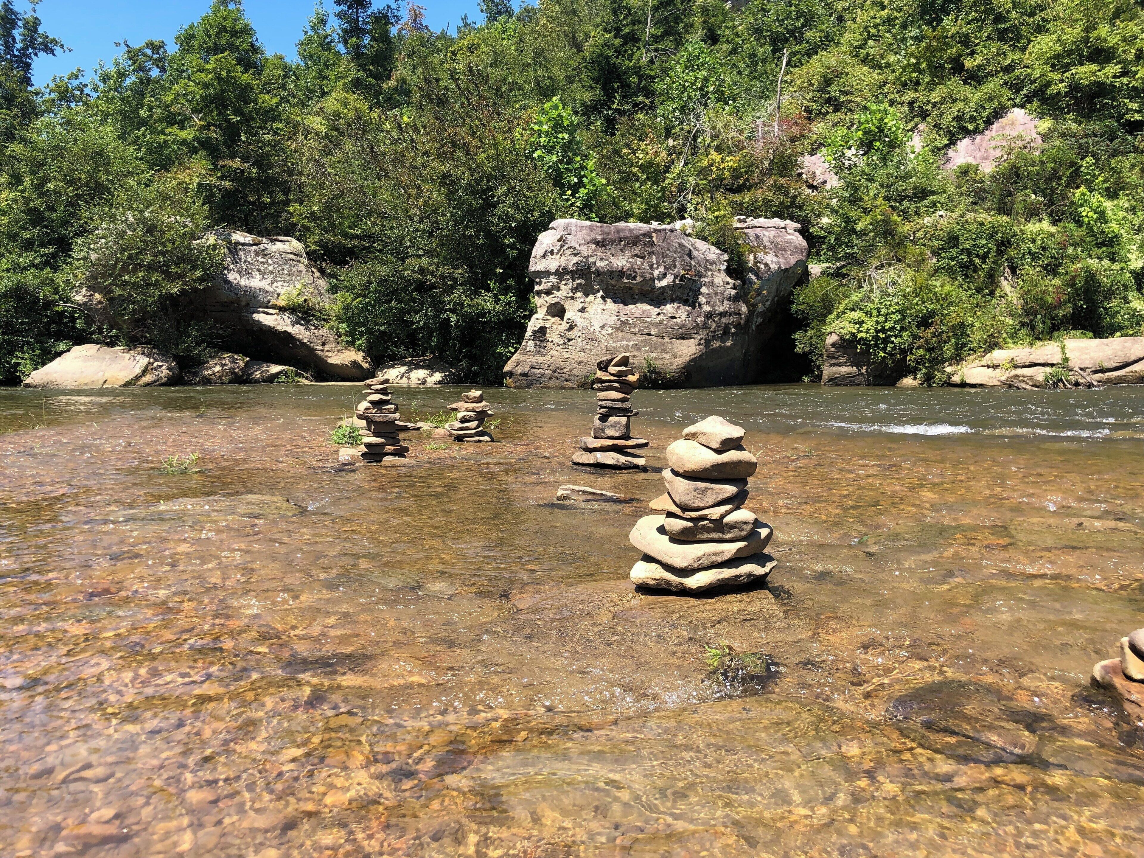 A clear river with stacks of rocks in the foreground and a forested hillside in the background.