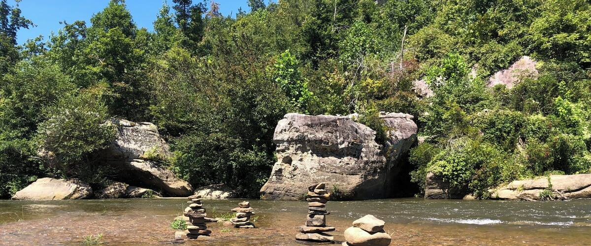 A clear river with stacks of rocks in the foreground and a forested hillside in the background.