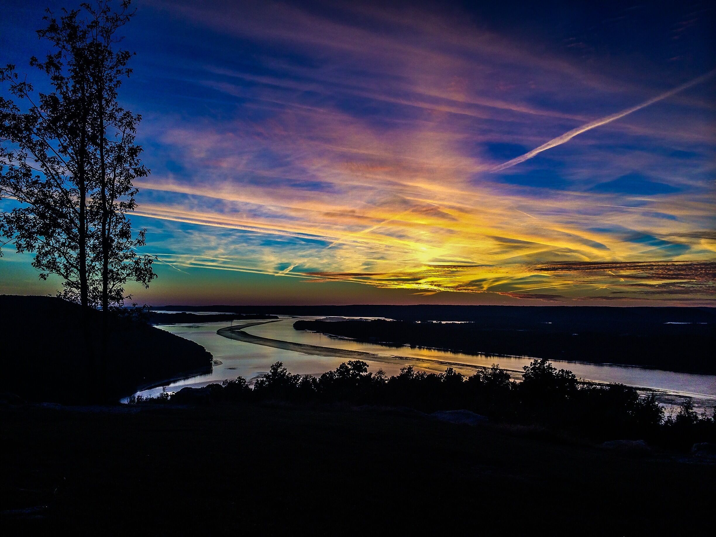 A river winding through a landscape with a tree on the left and a sunset with clouds in the sky.