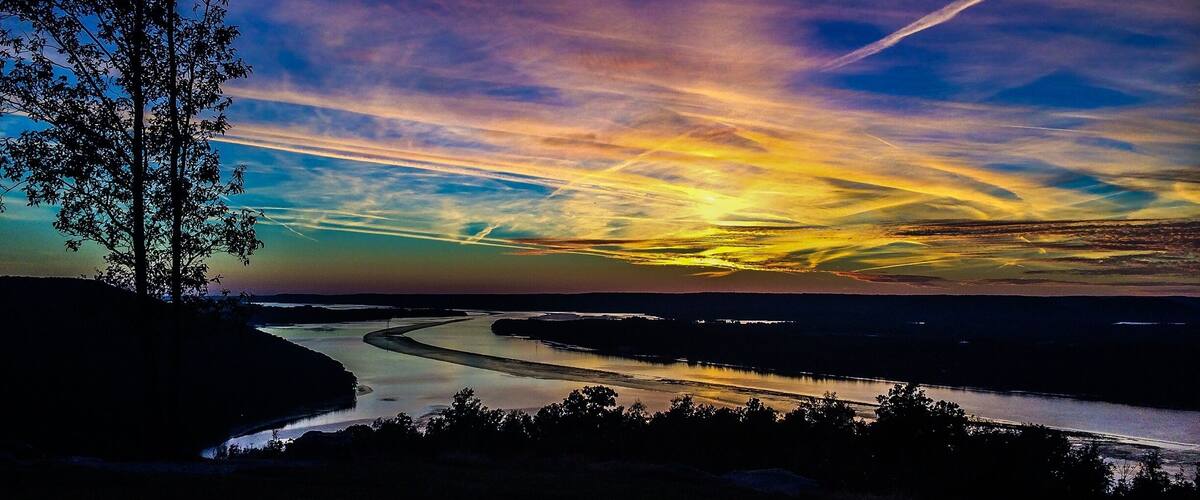 A river winding through a landscape with a tree on the left and a sunset with clouds in the sky.