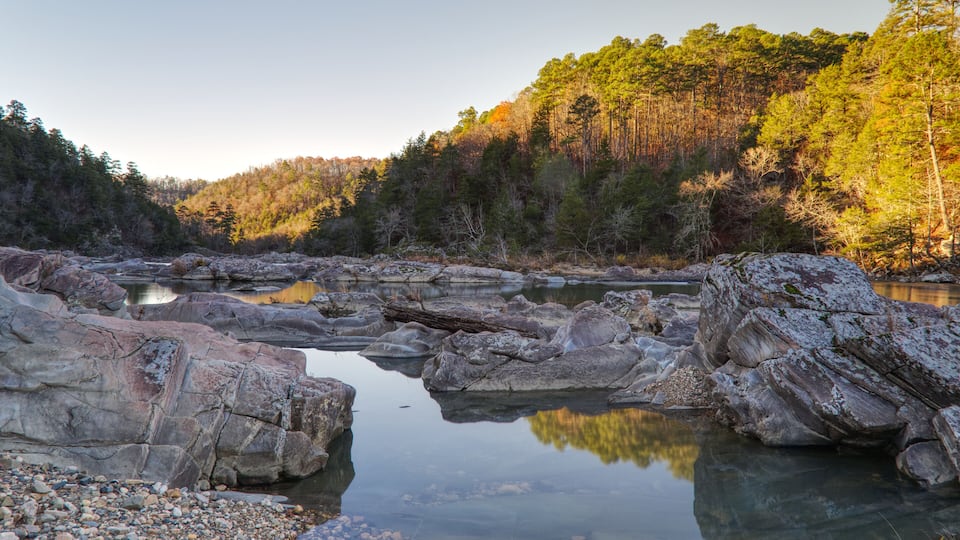 A calm river with large rocks in the foreground and a forested hillside in the background.