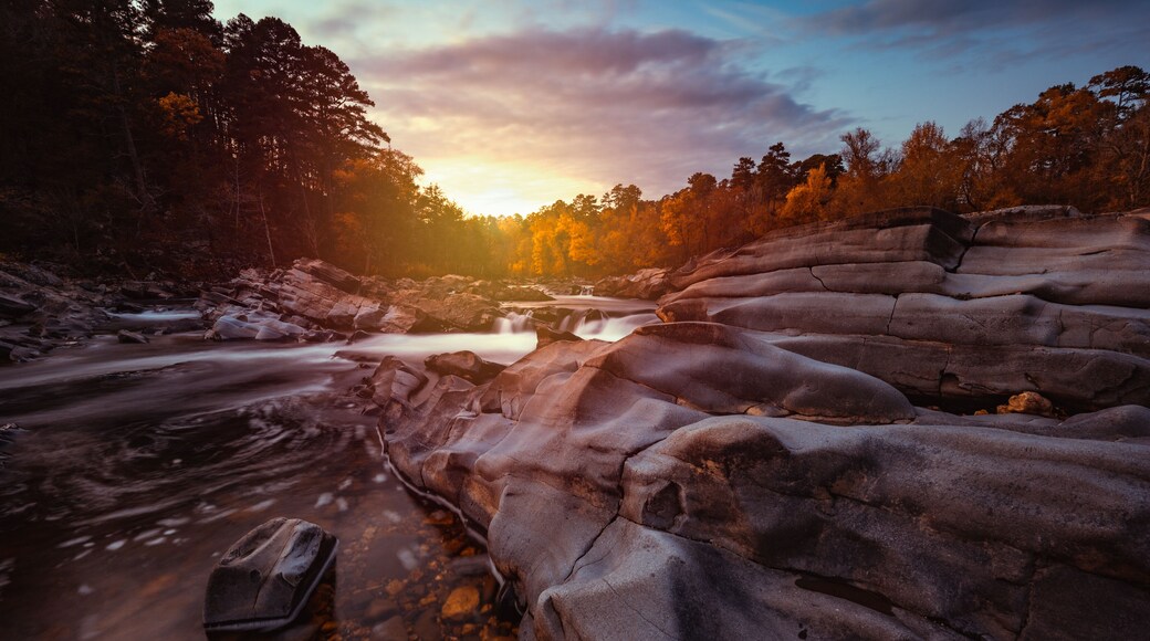 Cossatot Falls State Park at sunset during autumn in Arkansas as the water flows and cascades over smooth boulder rocks with colorful clouds above.