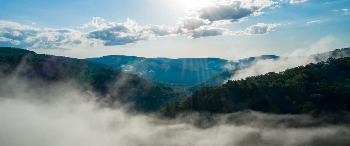 Paisaje montañoso con un valle cubierto de niebla, rayos de sol abriéndose paso entre las nubes y un cielo azul despejado.