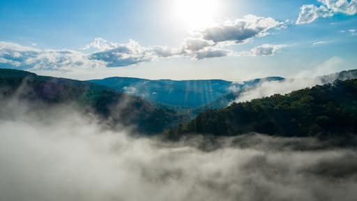 Paisaje montañoso con un valle cubierto de niebla, rayos de sol abriéndose paso entre las nubes y un cielo azul despejado.
