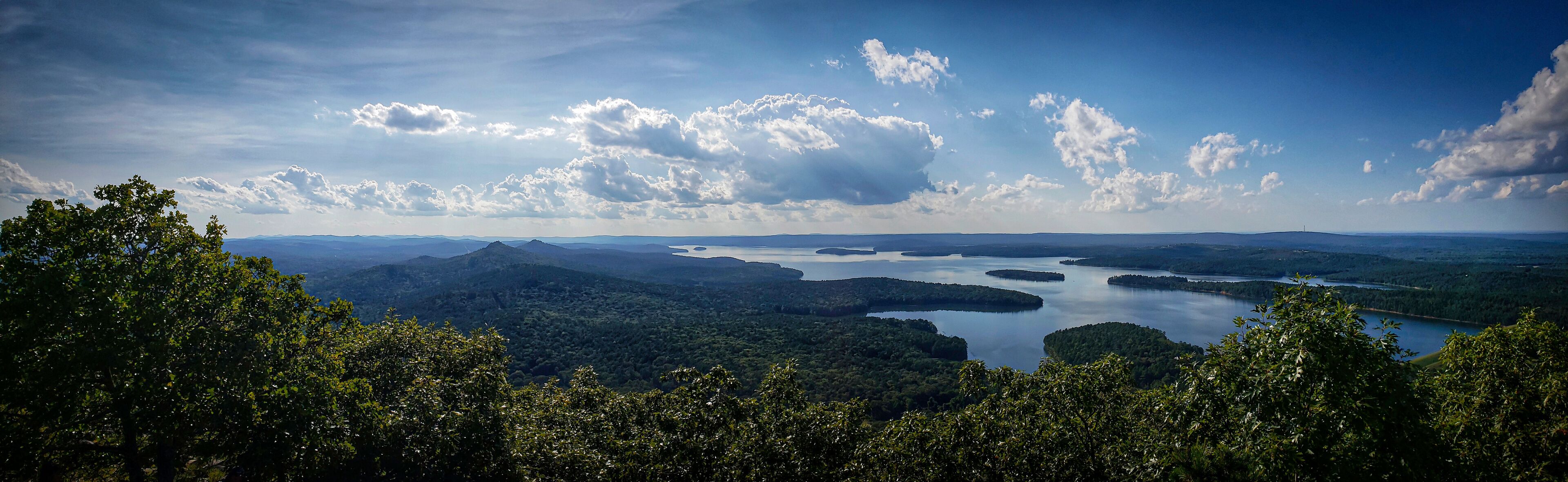 A mountainous landscape with a lake surrounded by dense forests and a clear sky with scattered clouds.