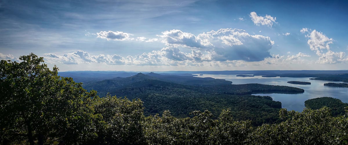 A mountainous landscape with a lake surrounded by dense forests and a clear sky with scattered clouds.