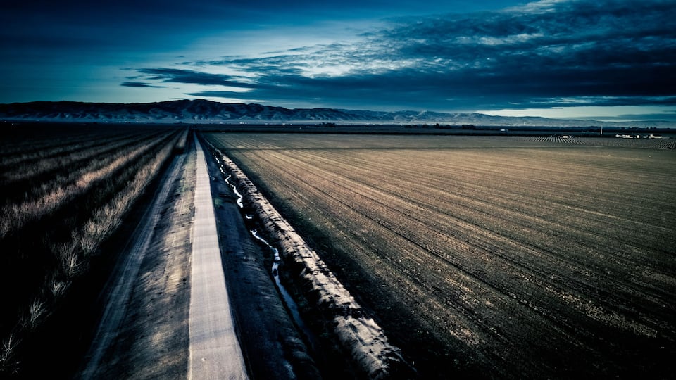 A straight road cutting through a vast field with distant mountains under a cloudy sky.