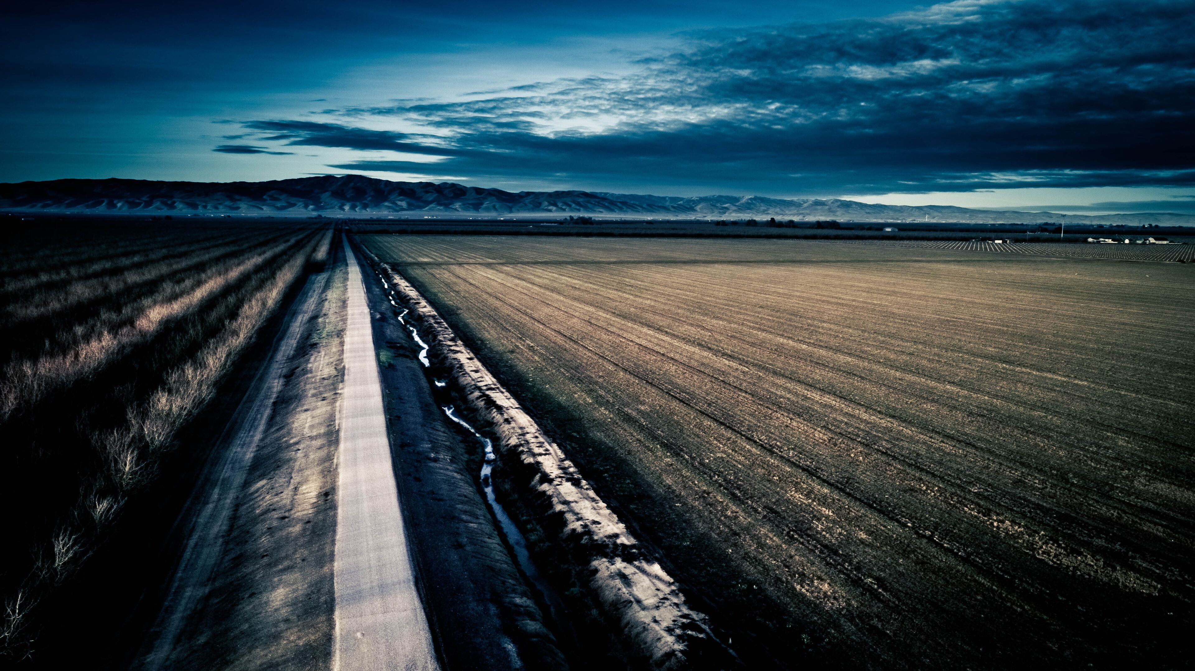 A straight road cutting through a vast field with distant mountains under a cloudy sky.
