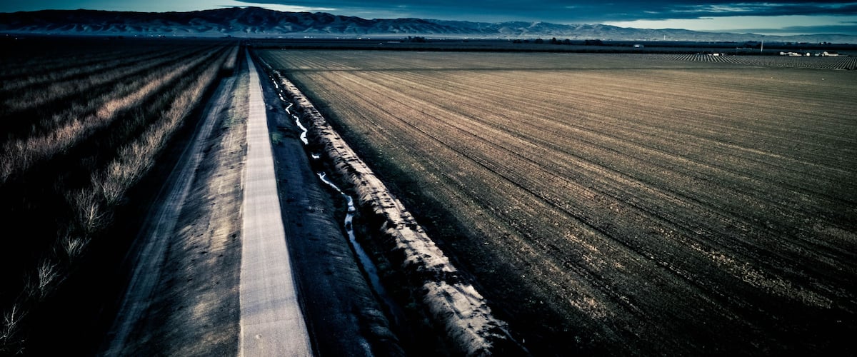 A straight road cutting through a vast field with distant mountains under a cloudy sky.