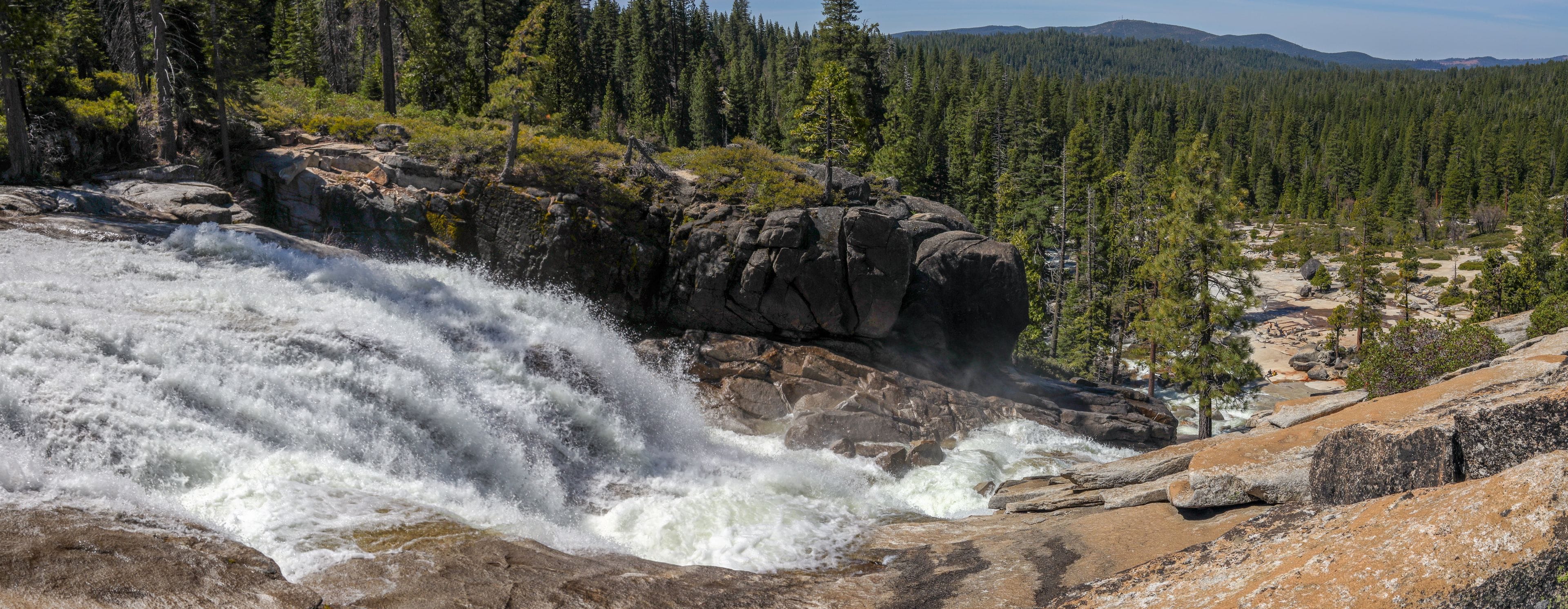 Une cascade d’eau se précipitant sur des rochers, entourée d’une forêt dense et de montagnes lointaines.