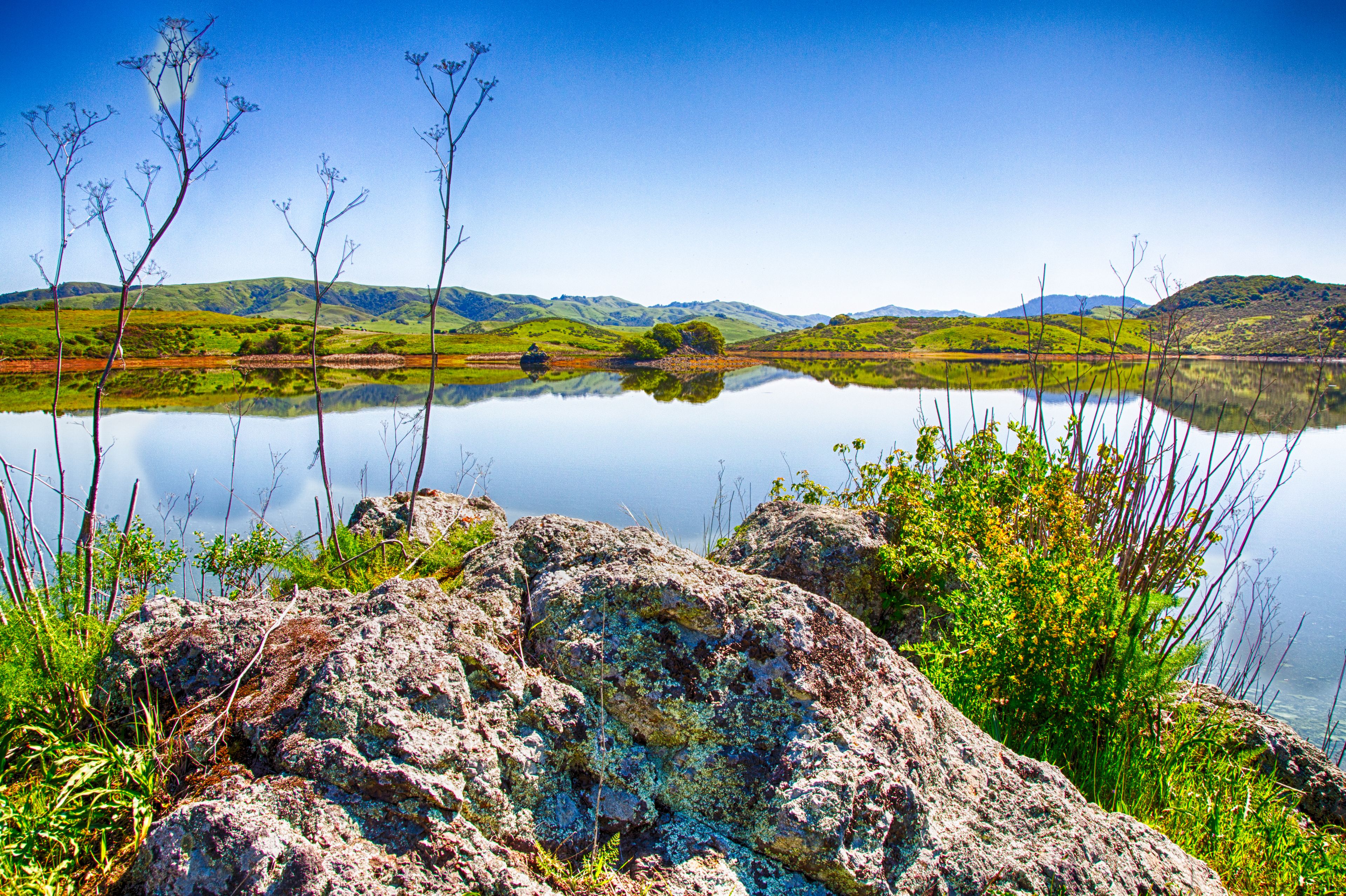 A calm lake with clear reflections, surrounded by green hills and rocky outcrops.