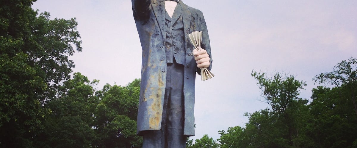 A statue of a man in a suit holding a fan, standing on a pedestal in a park with trees in the background.