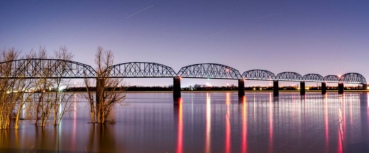 A steel bridge over calm water at dusk with star trails in the sky.