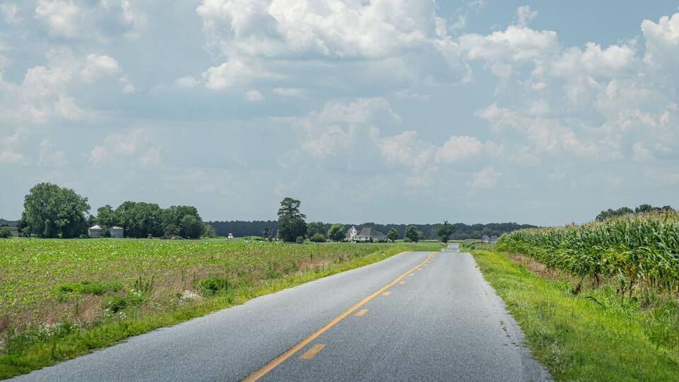 A rural road flanked by fields and trees under a partly cloudy sky.