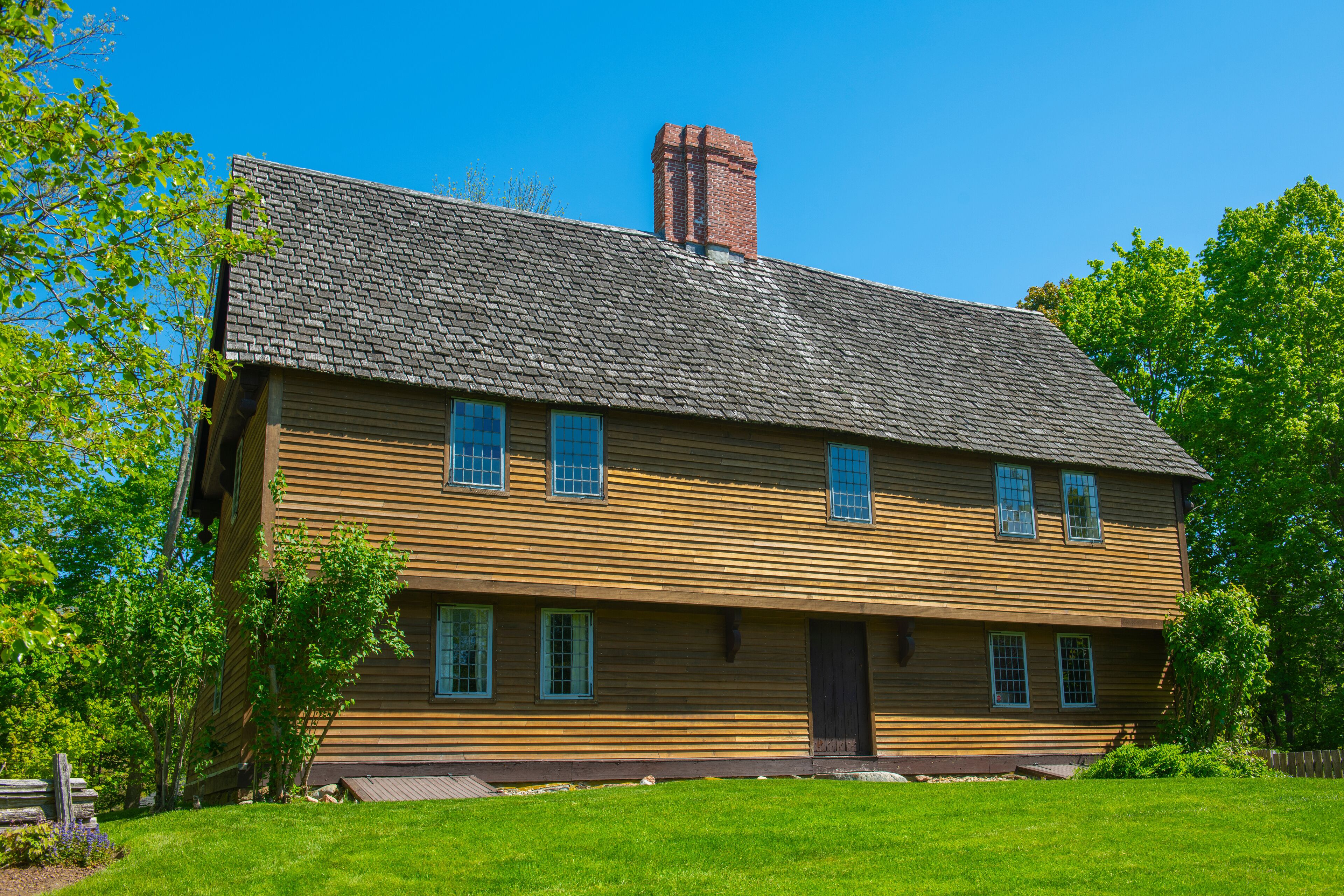 A large wooden house with a steep roof and a brick chimney, surrounded by green trees and a well-maintained lawn.
