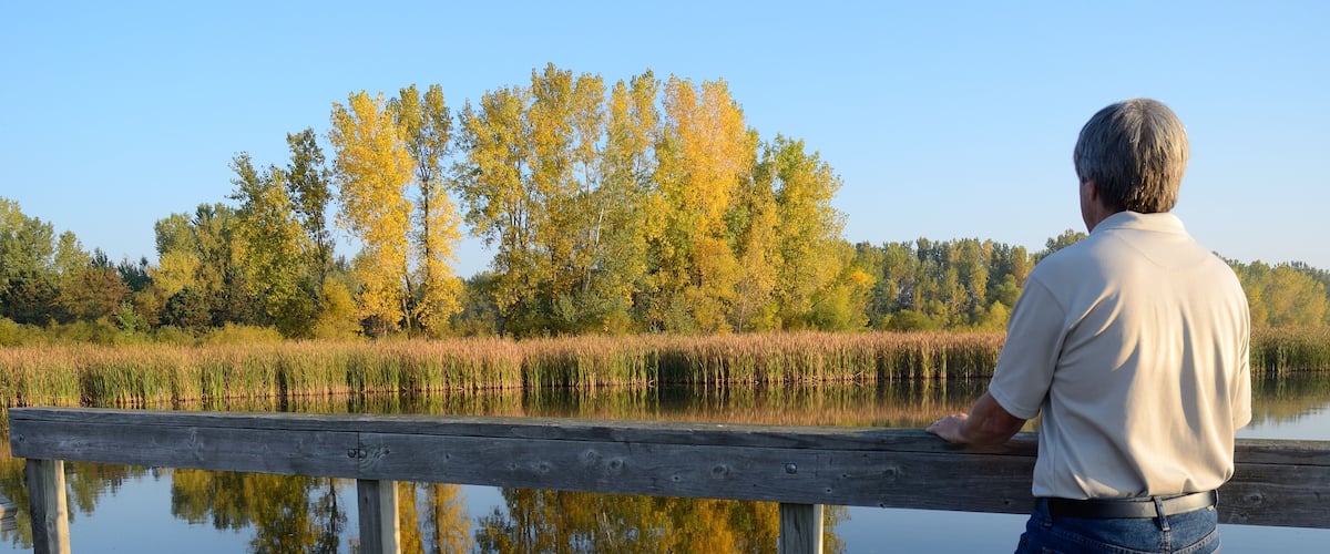 A man standing on a wooden dock overlooking a calm lake with autumn-colored trees in the background.