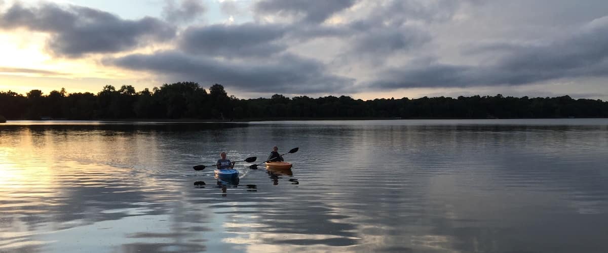 Two kayakers on a calm lake with a forested shoreline and a cloudy sky.