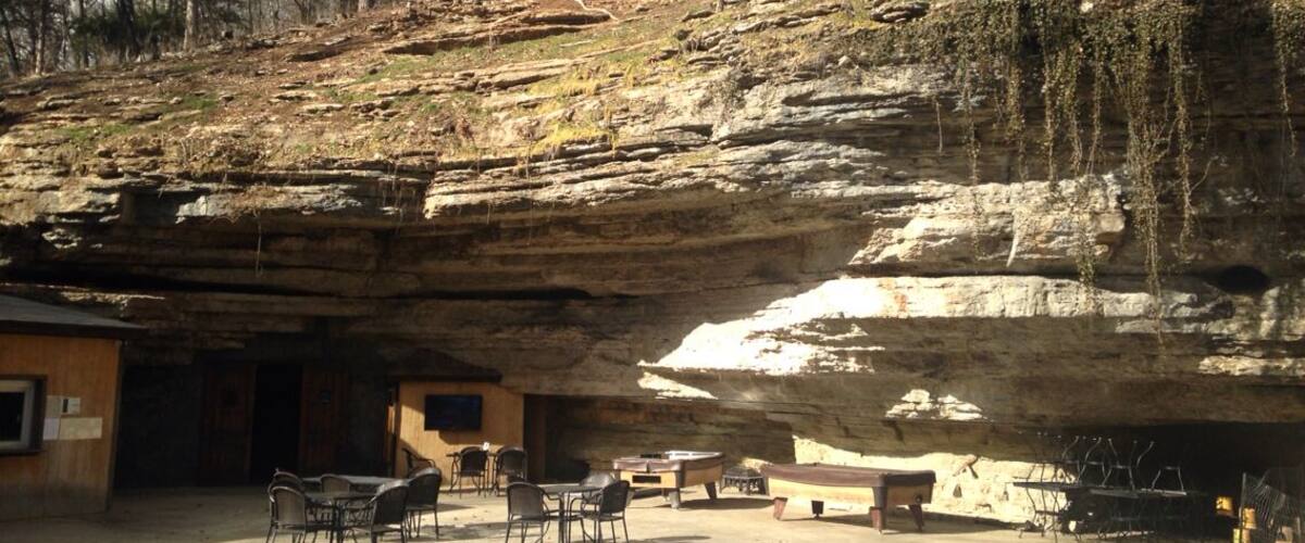 A natural rock formation with a cave entrance, outdoor seating, and a forested background.
