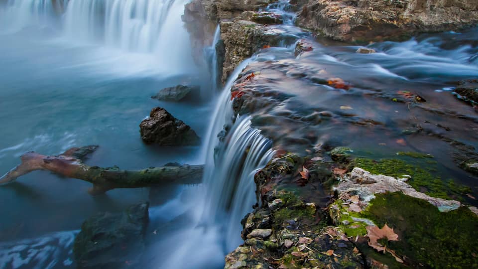 A waterfall cascading over rocks with moss and fallen leaves.