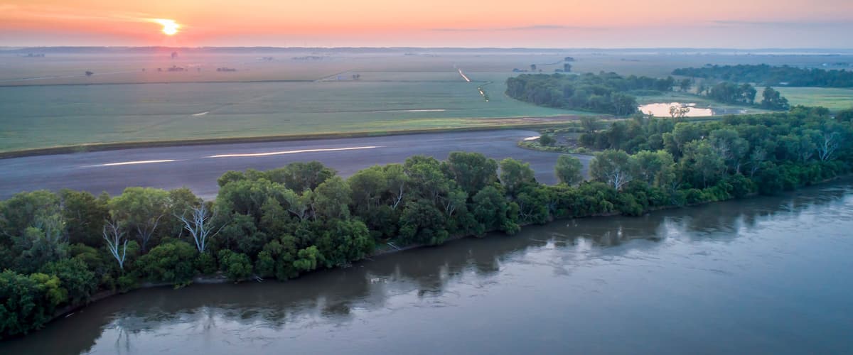 A riverbank with dense greenery, a runway, and a sunset over a flat landscape.