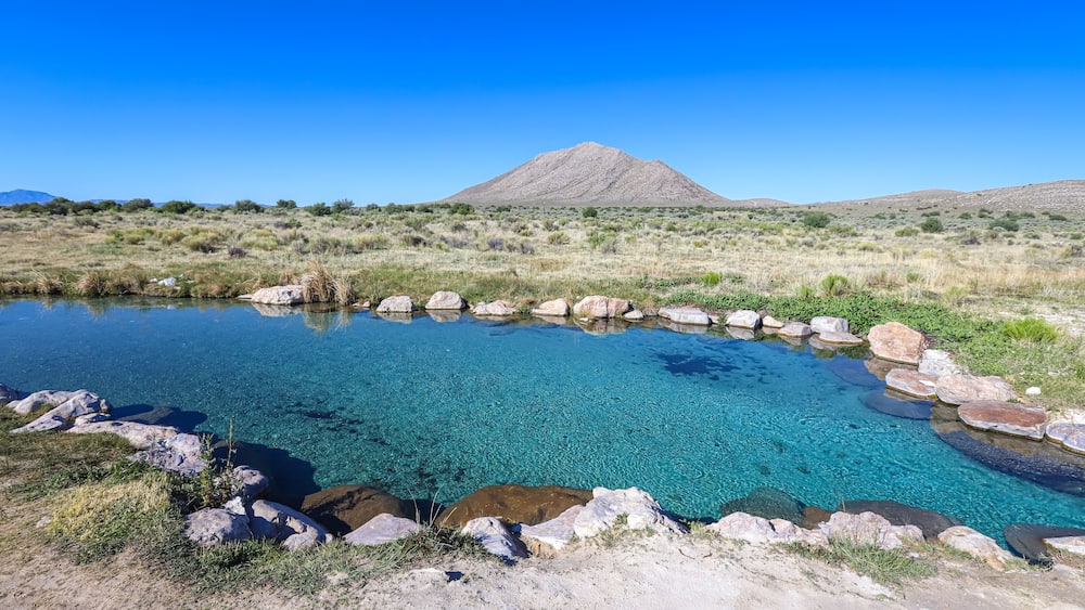 Ein klarer, blauer Teich, umgeben von Felsen, mit einem Berg im Hintergrund.