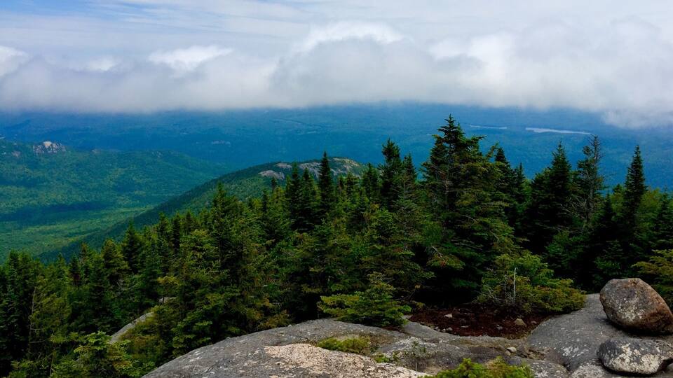 A mountainous landscape with dense coniferous forests, rocky outcrops, and a distant view of a lake under a partly cloudy sky.