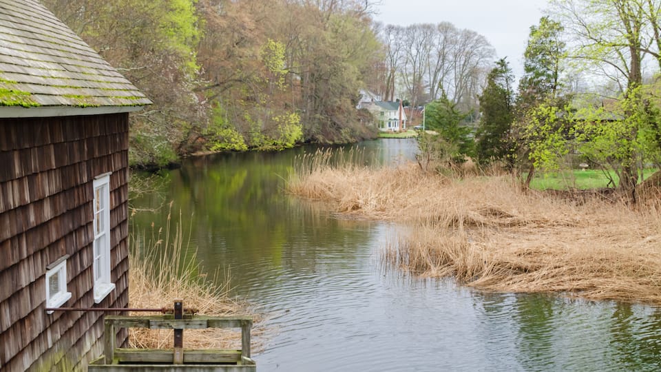 A wooden cabin by a calm river with a house in the background.