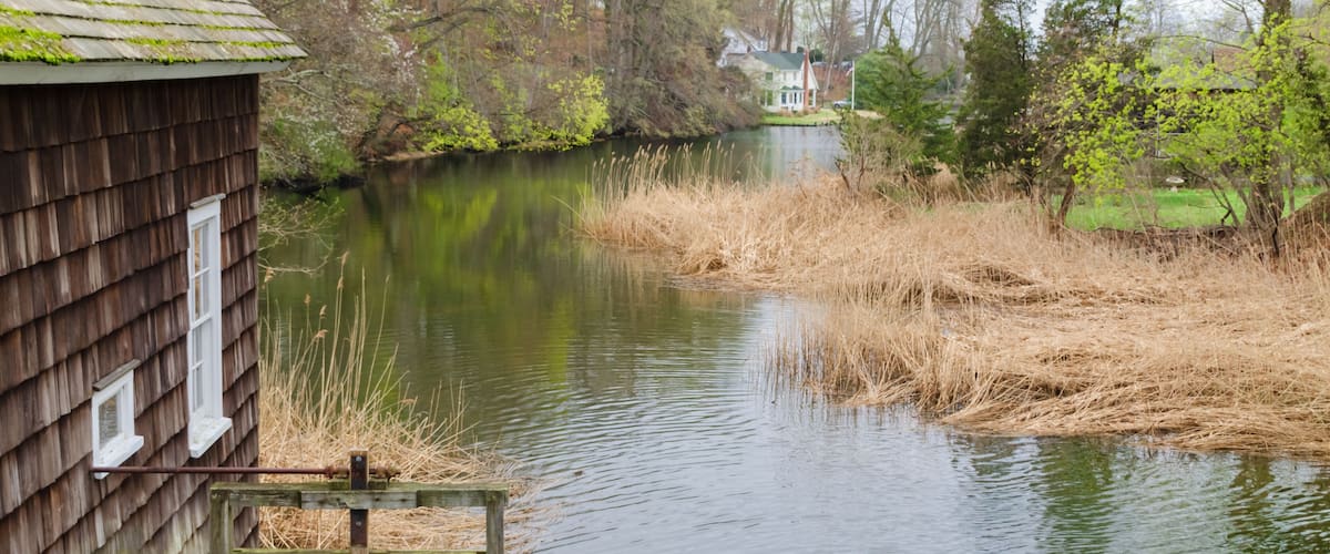 A wooden cabin by a calm river with a house in the background.