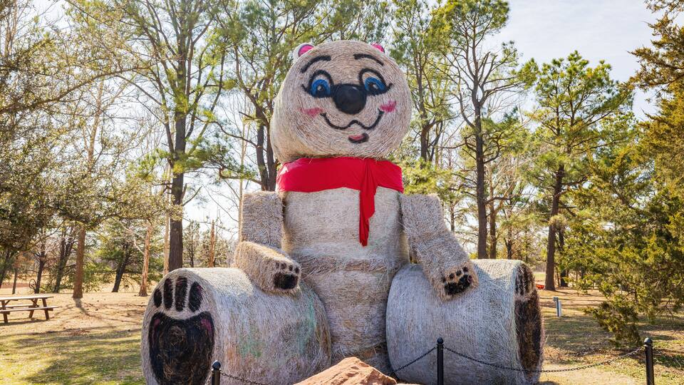 A large bear sculpture made of hay bales in a park with trees and picnic tables.