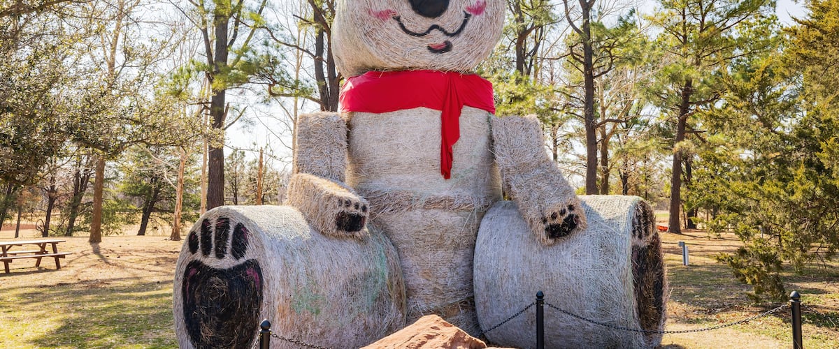 A large bear sculpture made of hay bales in a park with trees and picnic tables.