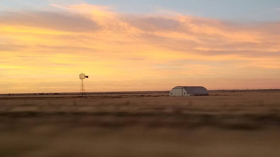 A windmill and a barn in a flat landscape during sunset.
