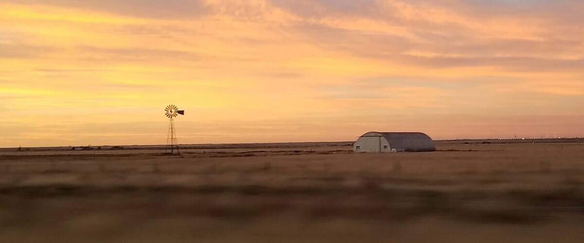 A windmill and a barn in a flat landscape during sunset.