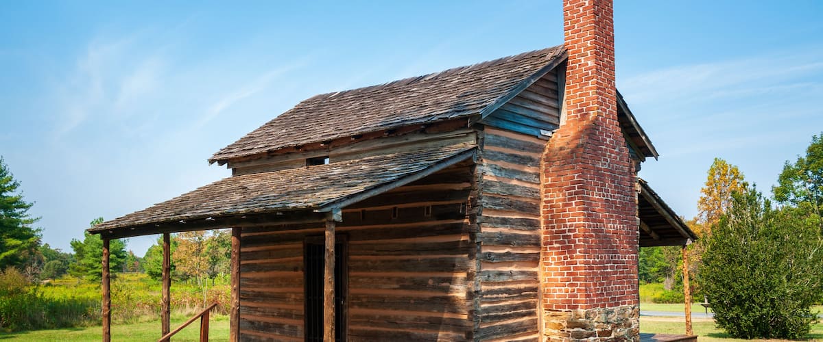 A rustic log cabin with a chimney, surrounded by lush greenery.