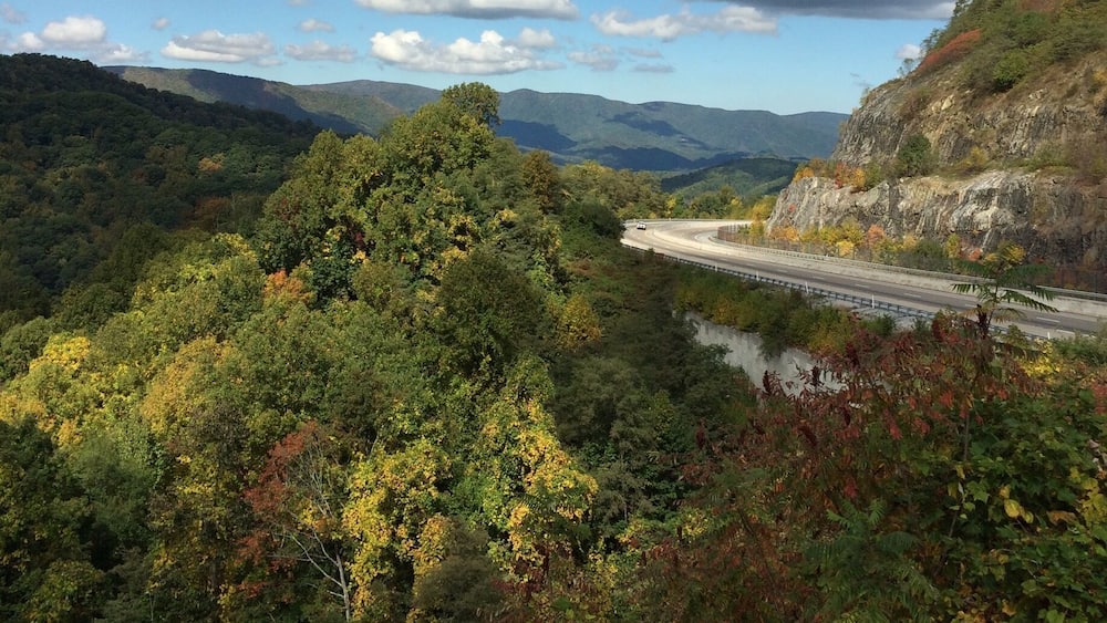 A winding road through a forested valley with a river and distant mountains under a partly cloudy sky.