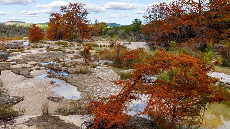 A natural landscape with a rocky stream, autumn-colored trees, and distant hills.
