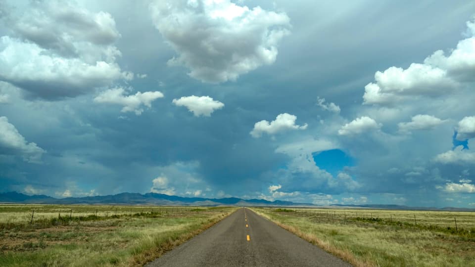 A straight road in a flat landscape with mountains in the distance and a cloudy sky.