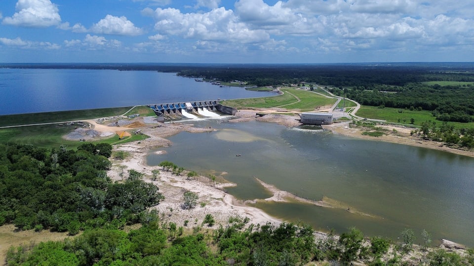 A dam with water flowing over it, a large reservoir, and surrounding greenery.