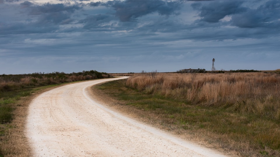 A dirt road curving through a grassy landscape with a windmill in the distance under a cloudy sky.