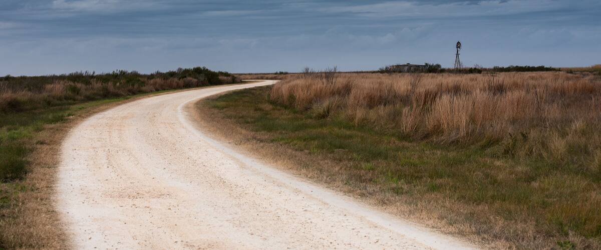 A dirt road curving through a grassy landscape with a windmill in the distance under a cloudy sky.