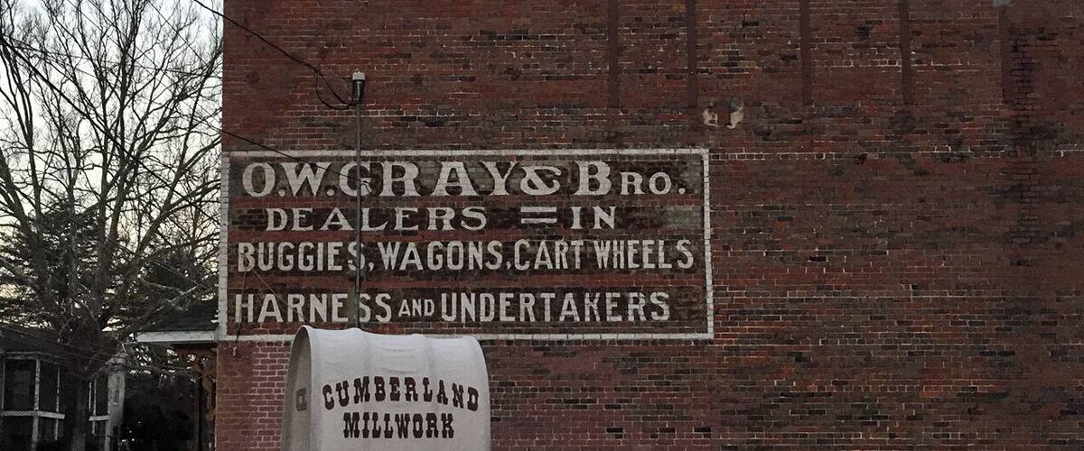 A brick building with a sign advertising O.W. Gray & Bro. Dealers in Buggies, Wagons, Cart Wheels, Harness and Undertakers. A truck labeled "Cumberland Millwork" is parked in front.
