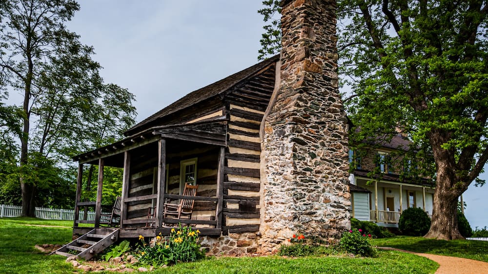 A rustic log cabin with a stone chimney, surrounded by lush greenery and a white picket fence.