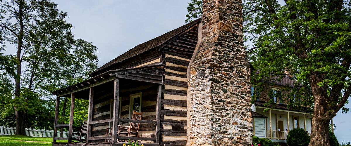 A rustic log cabin with a stone chimney, surrounded by lush greenery and a white picket fence.