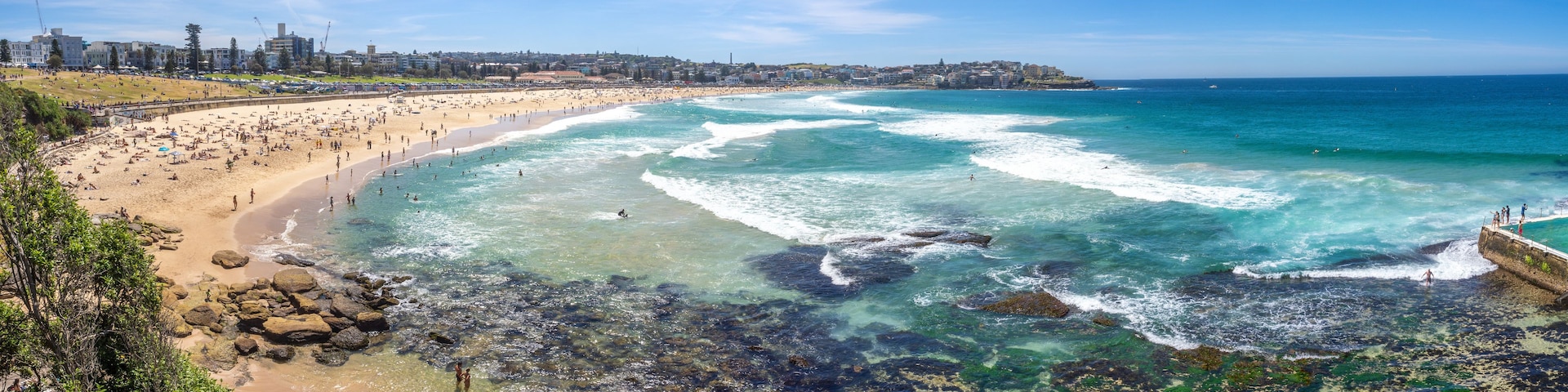 A coastal cityscape with a wide sandy beach, rocky shoreline, and a clear blue sky.