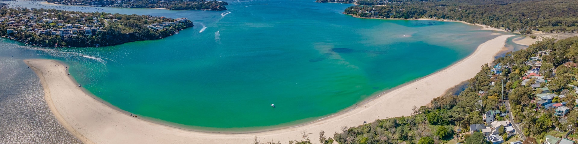 Vista aérea de uma área costeira com uma extensa praia arenosa, águas turquesa cristalinas e uma península densamente arborizada.