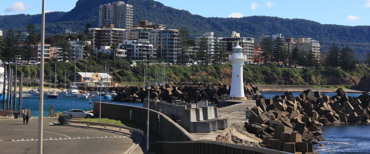 Un paisaje urbano costero con un faro, edificios residenciales y montañas al fondo.