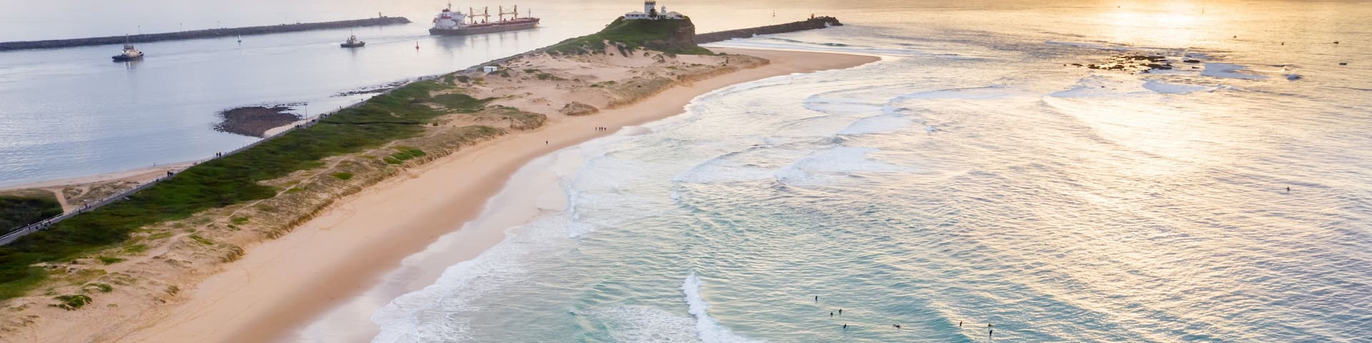 A coastal landscape with a sandy beach, ocean waves, a lighthouse, and cargo ships in the distance.