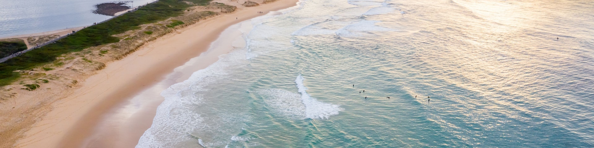 Paisagem costeira com uma praia de areia, ondas do oceano, um farol e navios cargueiros ao longe.