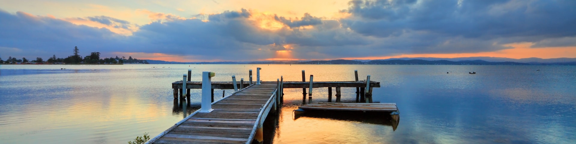 Eine Holzsteg streckt sich ins ruhige Wasser, im Hintergrund ein Sonnenuntergang.