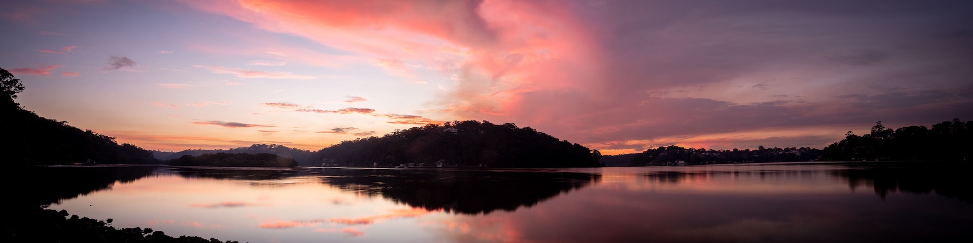 A calm lake reflecting a sunset sky with pink and purple hues, surrounded by hills and trees.