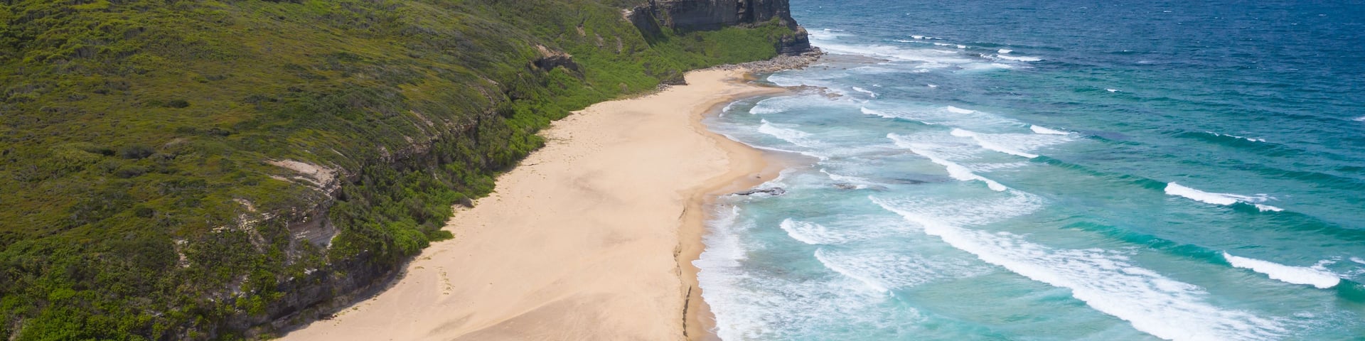 A coastal landscape with a sandy beach, green vegetation, and a cliff overlooking the ocean.